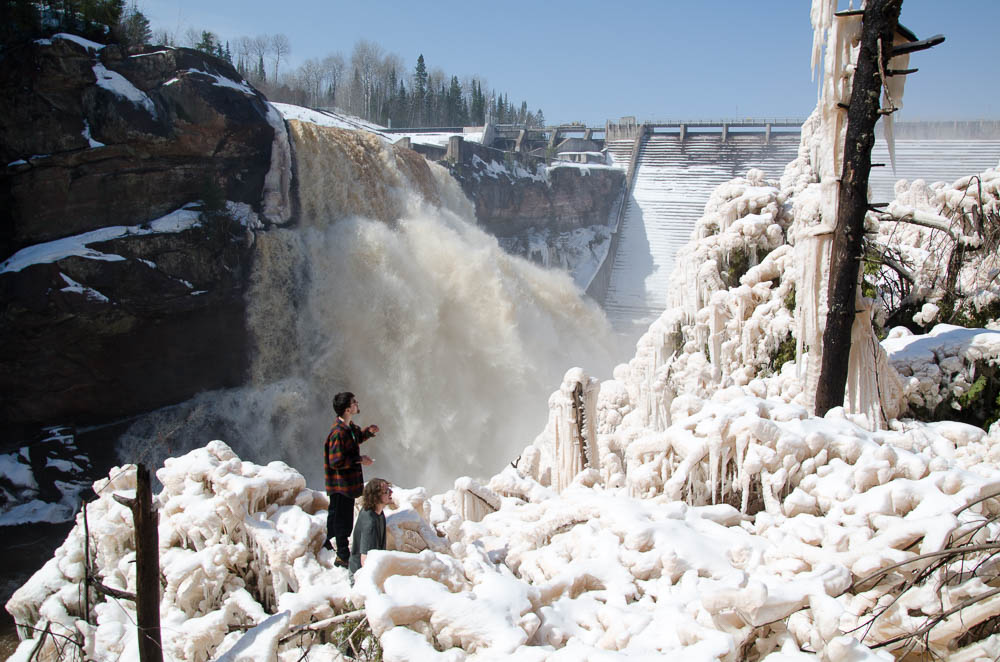 Two OVC members climbing over ice covered trees with a rushing spillway in the background